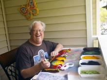 Todd LaFlame sitting at a table outside making a latch-hook rug.